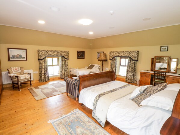 A bedroom with a large bed, a smaller bed, a desk, and a chair. There are two windows with curtains, a radiator, and a picture on the wall. - An Maide Bán (Cootehill)