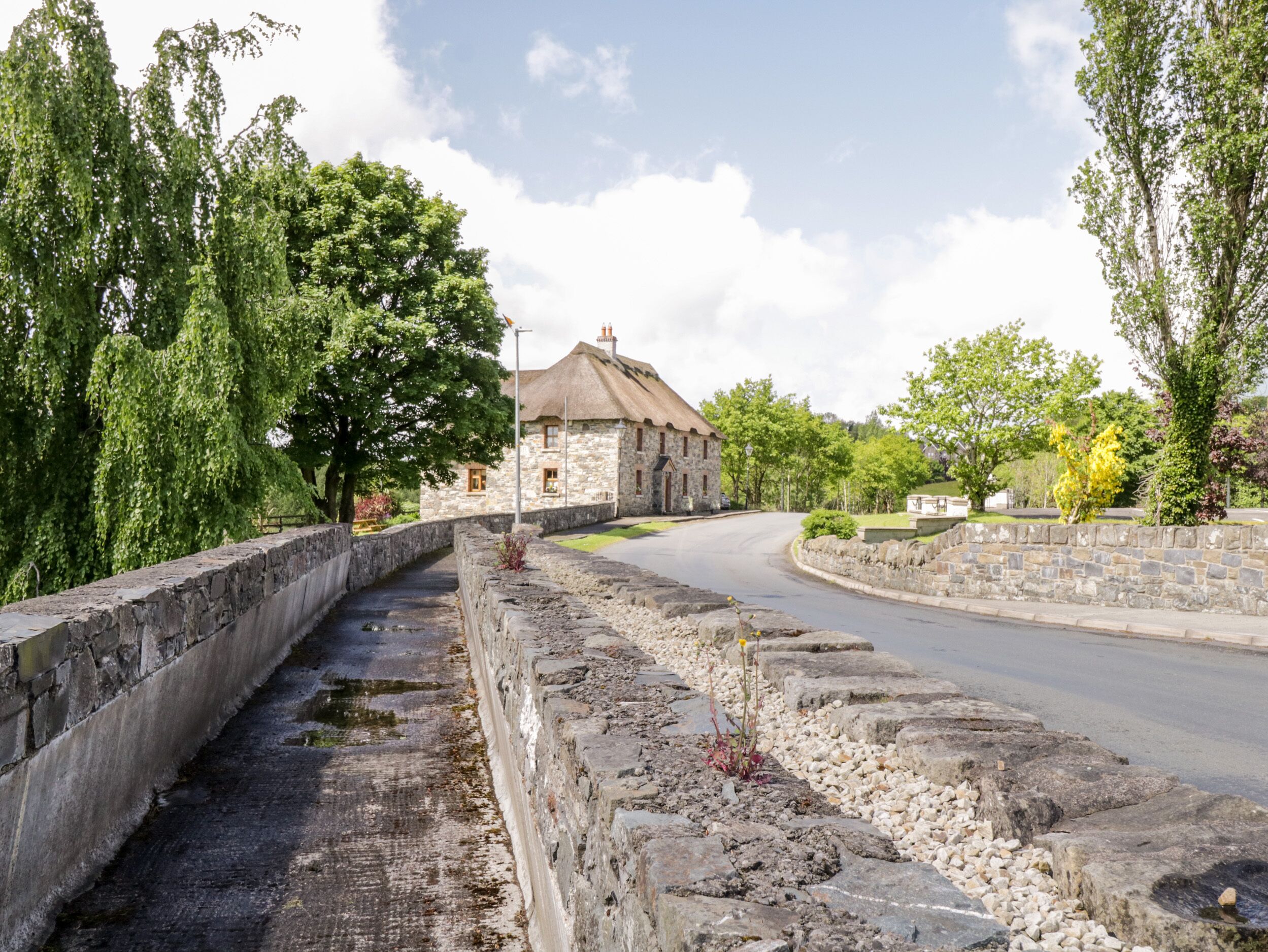 A stone bridge with a curved road, a stone building with a thatched roof, and lush greenery.