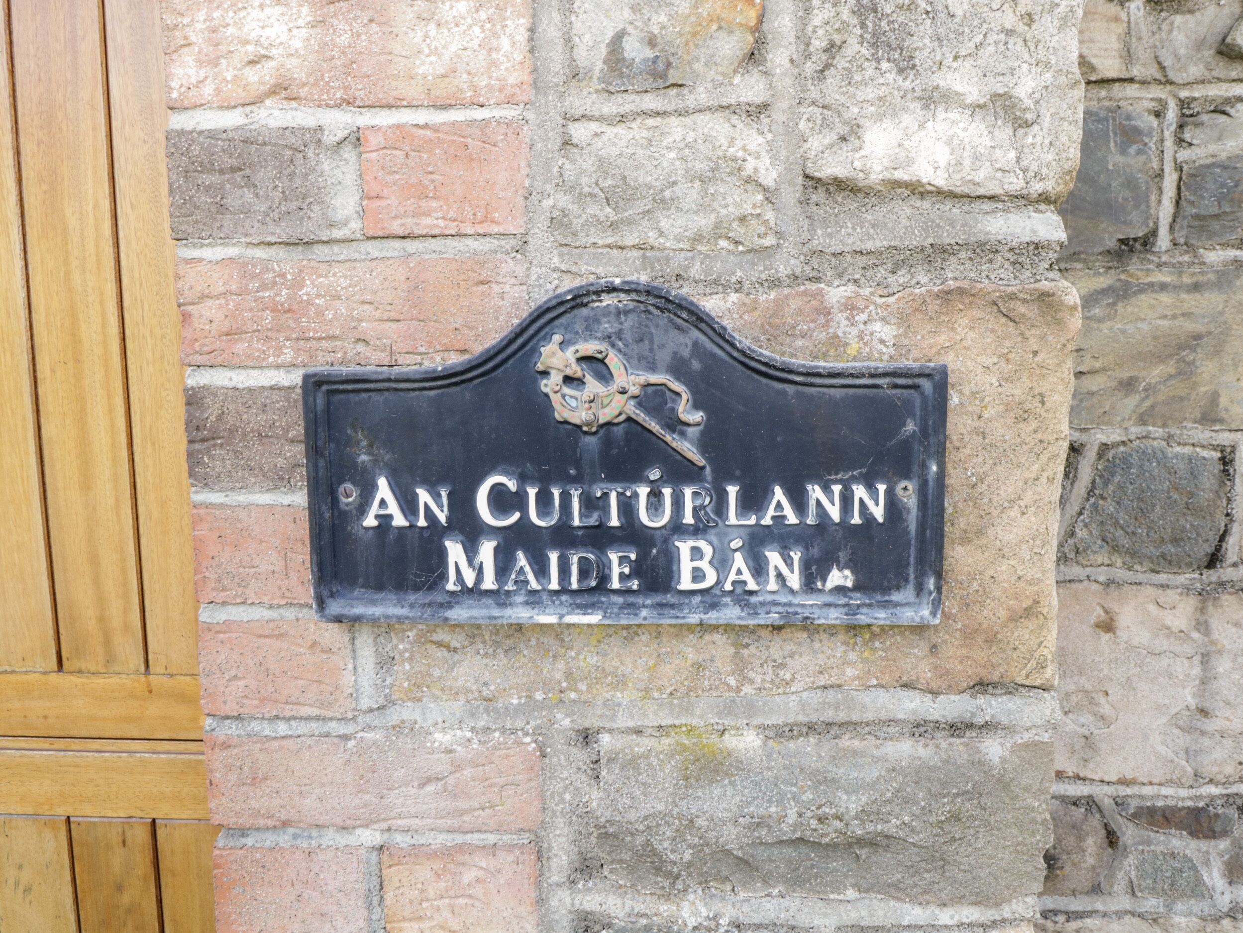 Une plaque en métal noir avec des lettres blanches, fixée à un mur de pierre, sur laquelle on peut lire : « AN CULTURLANN MAIDE BAN ».