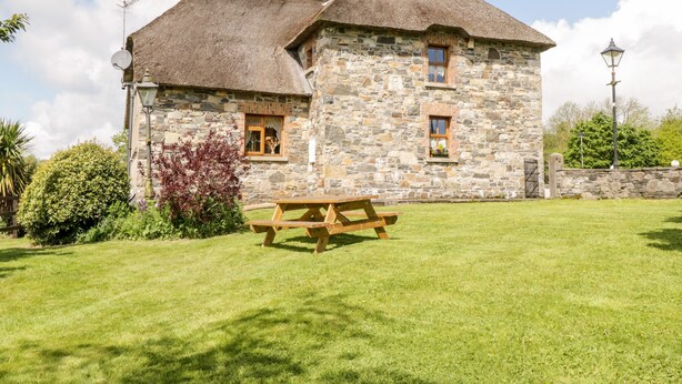 A stone building with a thatched roof, a picnic table, and a stone wall with a gate. - An Maide Bán (Cootehill)
