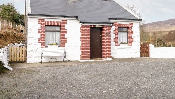 A small, single-story building with a white facade and red brick accents. It features a dark brown door, two windows with white frames, and a small porch area. - The Cottage (Newport)