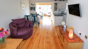 A living room with a purple sofa, wooden flooring, a dining area with chairs, a kitchen with white cabinets, and a TV mounted on the wall. - Teach Teolai (Carraroe)