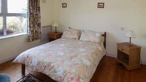 A bedroom with a bed, a nightstand, a lamp, a window with curtains, and a small wooden cabinet. - Number 12 (Mulranny)