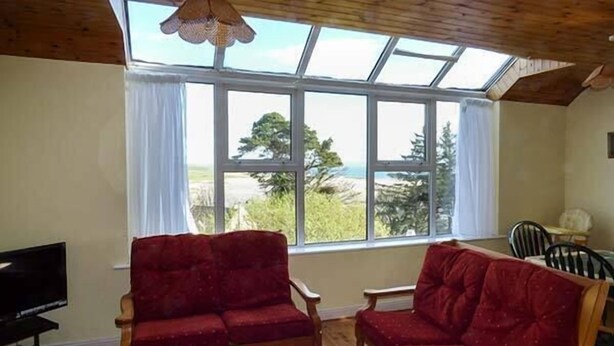 A living room with two red sofas, a wooden coffee table, and a TV. Large windows offer a view of the outdoors. - Number 12 (Mulranny)