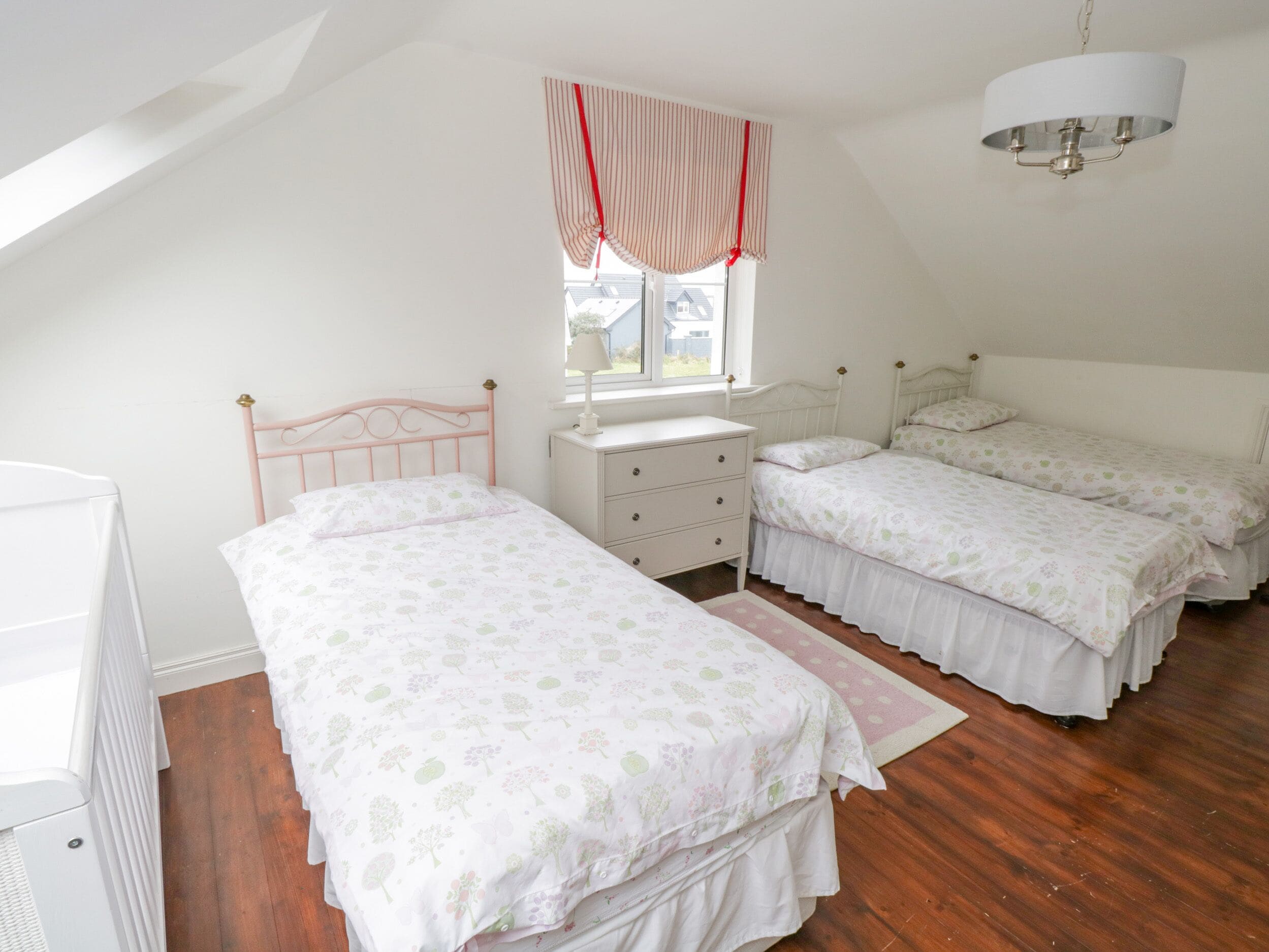 A bedroom with two single beds, a nightstand, a dresser, a window with curtains, and a ceiling light fixture.