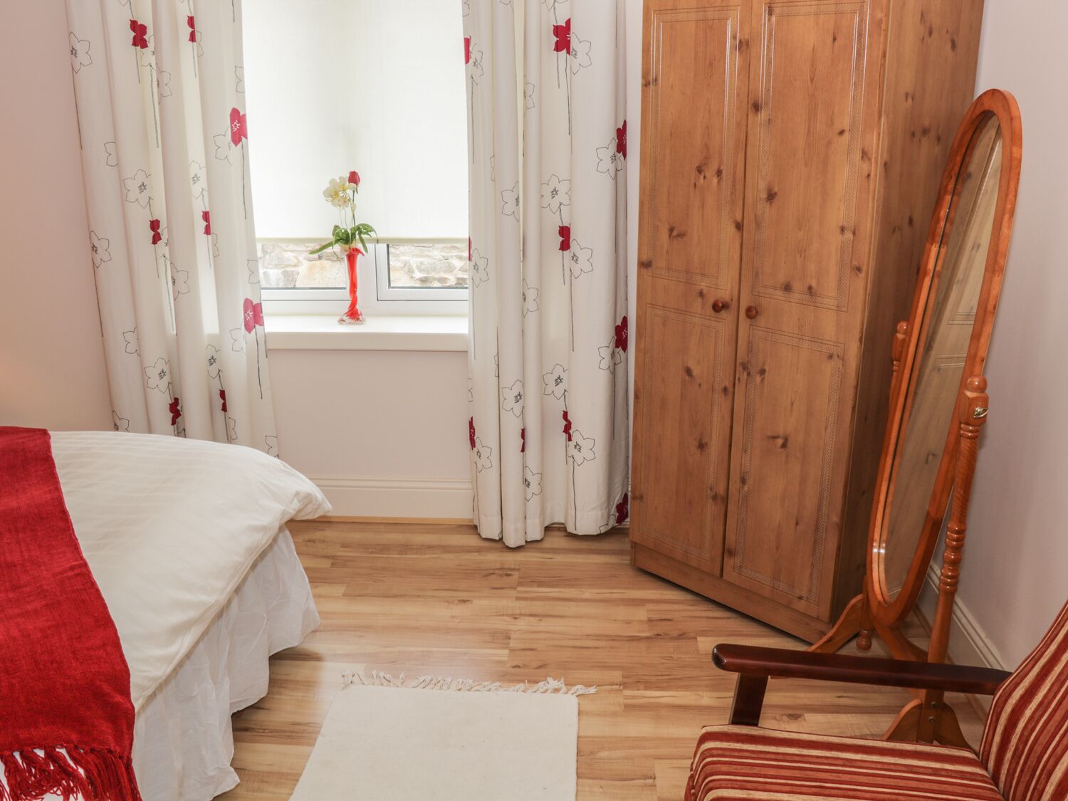A bedroom with a bed, a wooden wardrobe, a mirror, and a window with curtains.