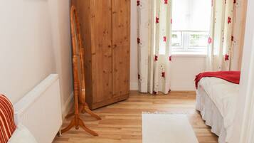 A bedroom with a wooden wardrobe, a wooden easel, a bed with white bedding, and a window with floral curtains.