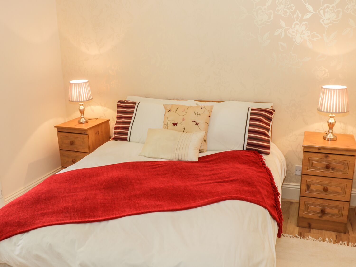 A neatly made bed with a red blanket, two bedside tables with lamps, and floral wallpaper.