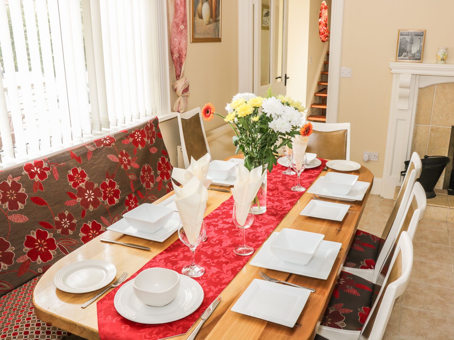Comedor con una larga mesa de madera dispuesta para cuatro, cojines rojos con estampado floral, platos blancos y un jarrón con flores.
