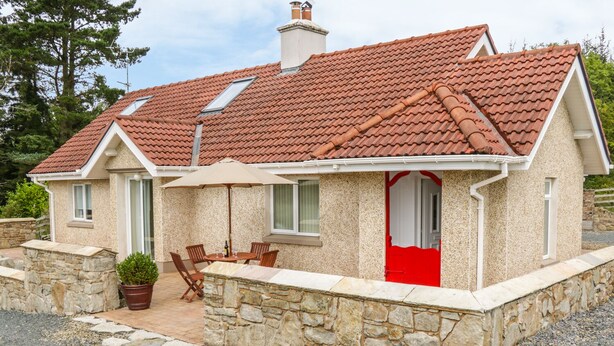 Une maison avec un toit recouvert de tuiles rouges, des murs en pierre, une porte rouge et une terrasse avec des chaises et une table.