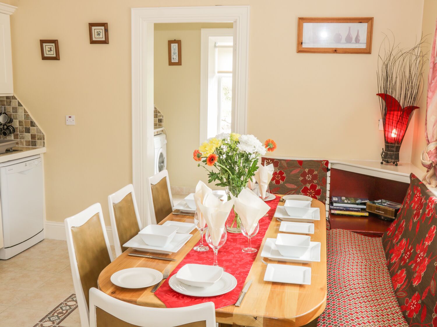 A dining area with a wooden table set for four, white chairs, and a red runner. A vase with flowers is on the table.