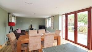 A dining area with a wooden table and chairs, a television, and a view of the outdoors through large sliding glass doors. - The Owl's Hoot (Tacumshane)