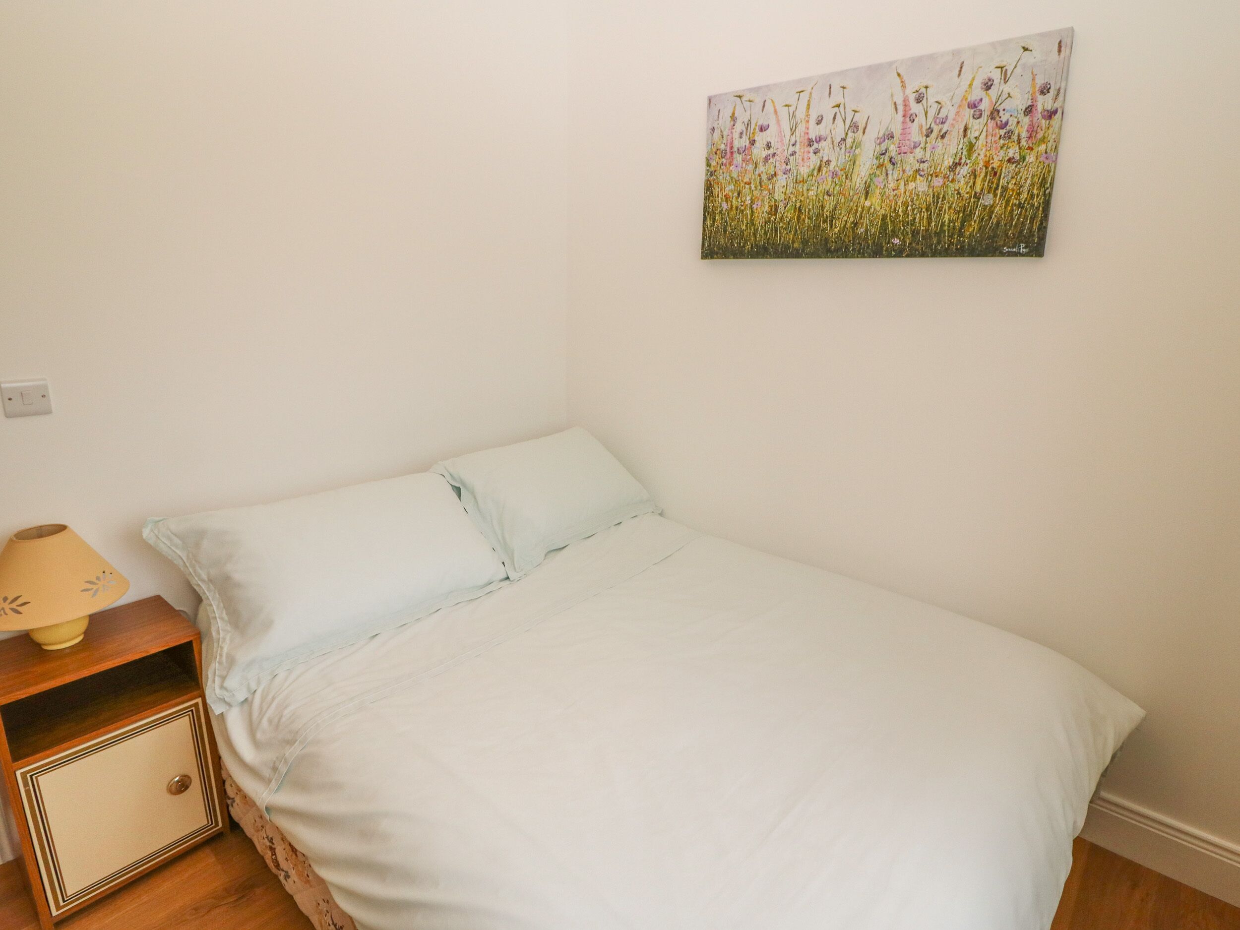 A single bed with a white sheet and a light blue pillow in a room with a white wall. There is a small wooden nightstand with a lamp and a drawer. A painting of flowers hangs on the wall above the bed.