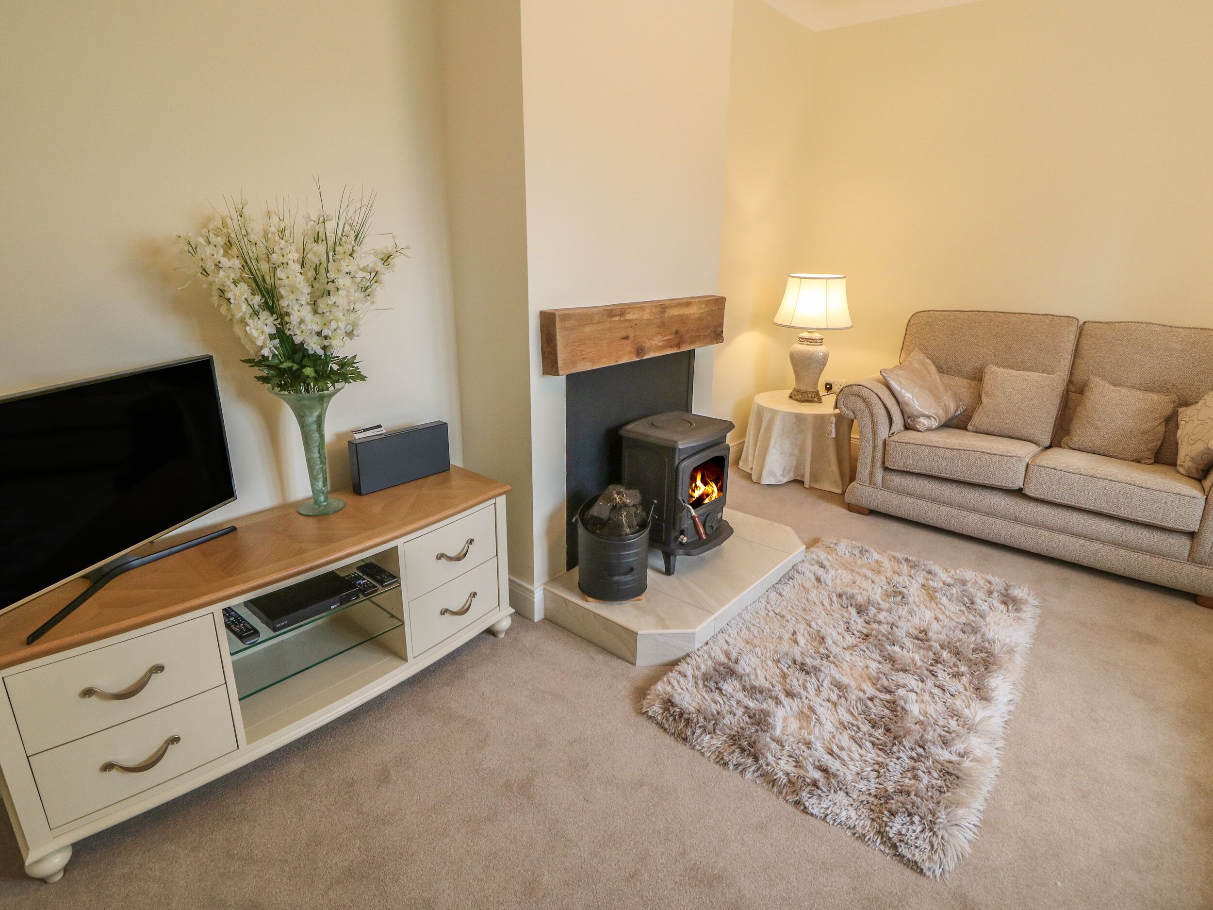 A living room with a flat-screen TV, a white cabinet with drawers, a small table with a lamp, a beige sofa, and a wood-burning stove.