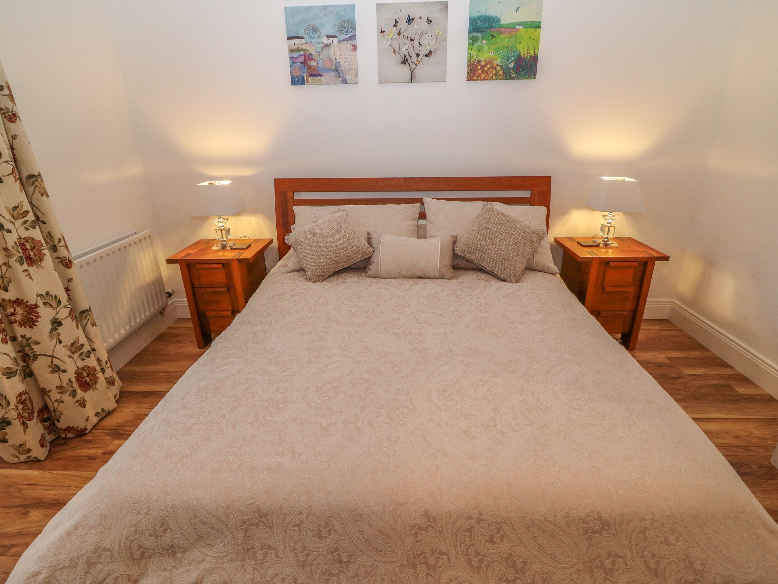 A neatly made bed with a patterned bedspread, flanked by two bedside tables with lamps, in a room with wooden flooring and a radiator.