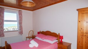 Un dormitorio con cama de madera, armario de madera, una silla, mesita de noche con lámpara y ventana con cortinas.