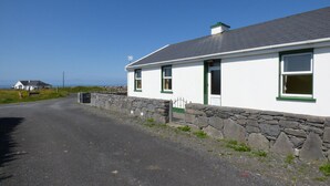 Un edificio blanco con detalles en verde y un muro de piedra como base, situado al lado de una carretera con vista despejada al mar.