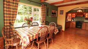 A dining area with a wooden table and chairs, a window with curtains, and a kitchen visible through an archway. - The Lodge (Beaufort)