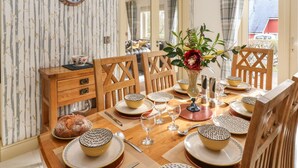 Een eetkamer met een houten tafel gedekt voor vier personen, een vaas met bloemen en een klok aan de muur.