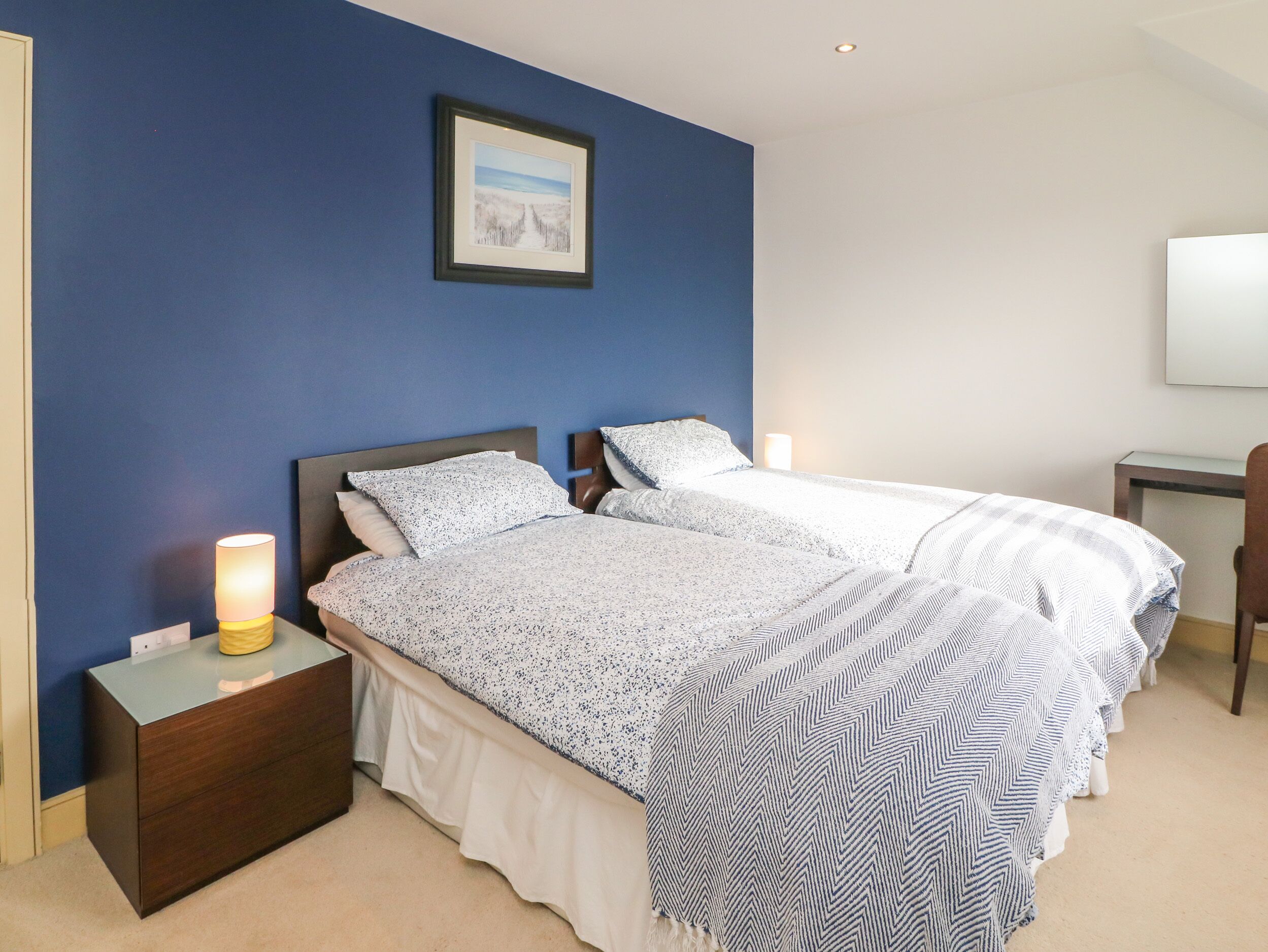 A double bed with a patterned bedspread, a nightstand with a lamp, and a framed picture on the wall.