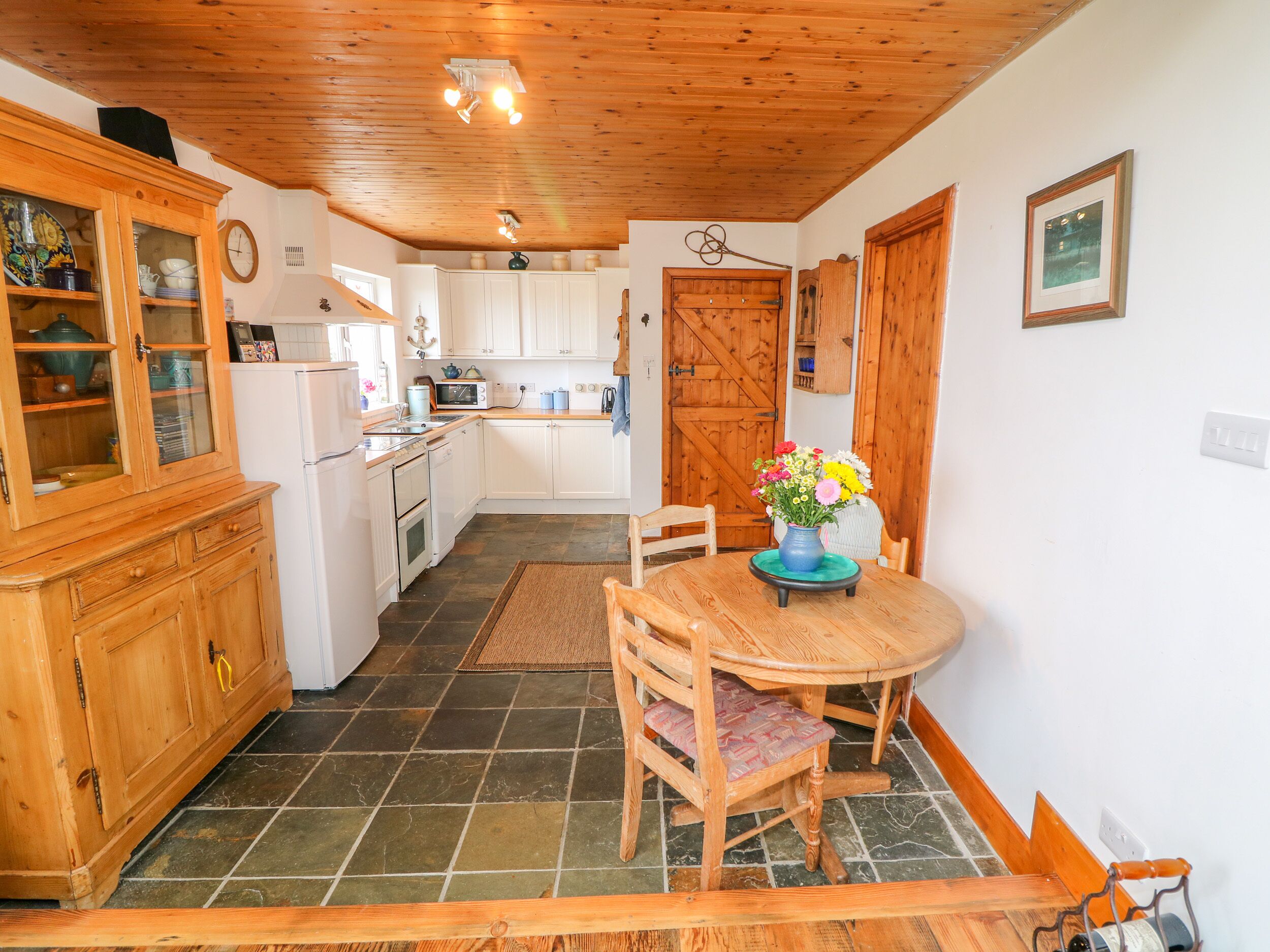 A kitchen with white cabinets, a wooden dining table with chairs, a vase of flowers, and a wooden ceiling.
