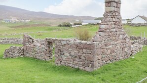 A stone ruin with a chimney, surrounded by a grassy field and a body of water in the background.
