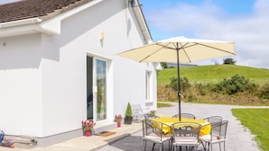 A white building with a patio area featuring a table and chairs under a large umbrella, a door, and potted plants. - Artigallivan (Killarney)