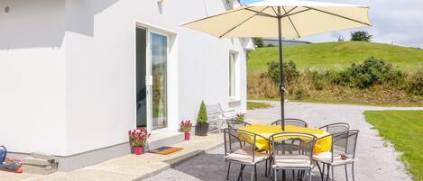 A white building with a patio area featuring a table and chairs under a large umbrella, a door, and potted plants.