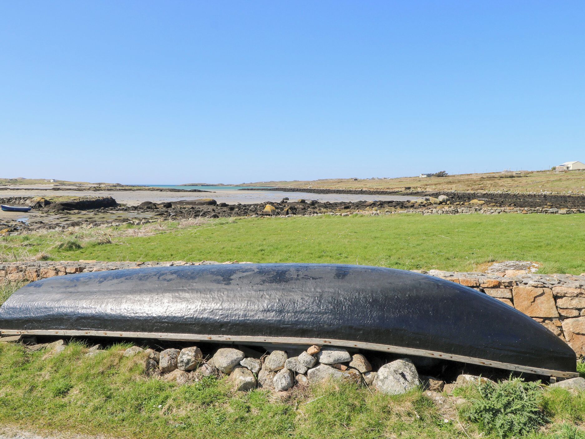 A large, black, curved object on a grassy field with a stone wall in the foreground and a coastal landscape in the background.
