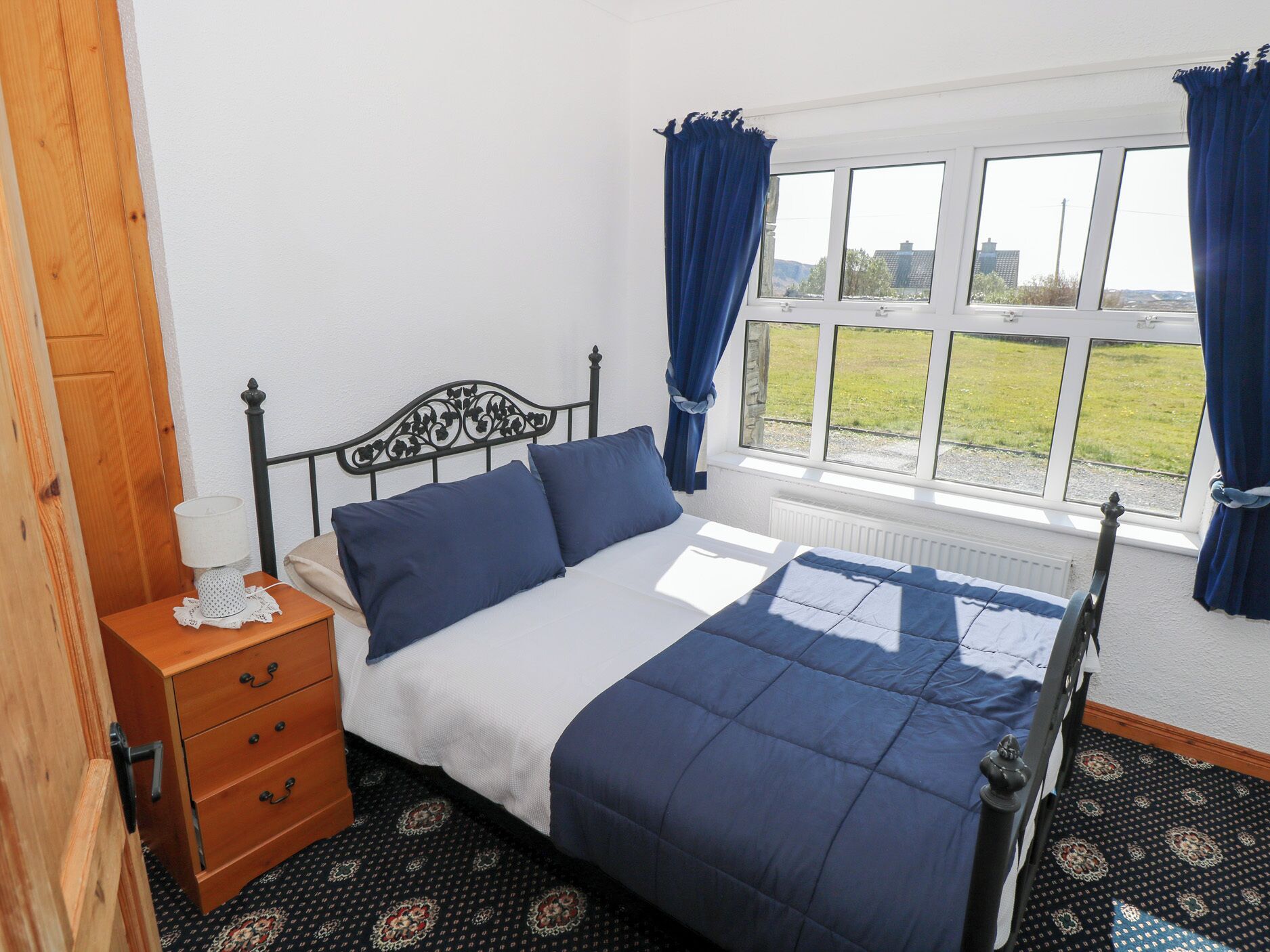 A bedroom with a bed, a wooden nightstand, a window with curtains, and a view of the outdoors.