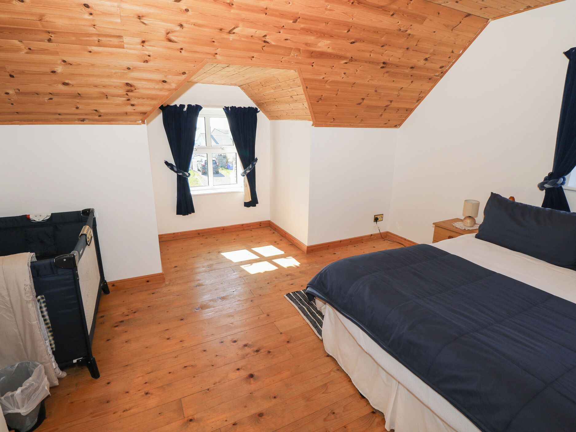 Quarto com teto inclinado, piso de madeira, cama com roupa de cama escura, janela com cortinas e pequena mesa de cabeceira com abajur.