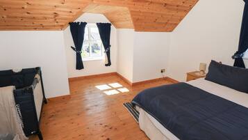 Quarto com teto inclinado, piso de madeira, cama com roupa de cama escura, janela com cortinas e pequena mesa de cabeceira com abajur.