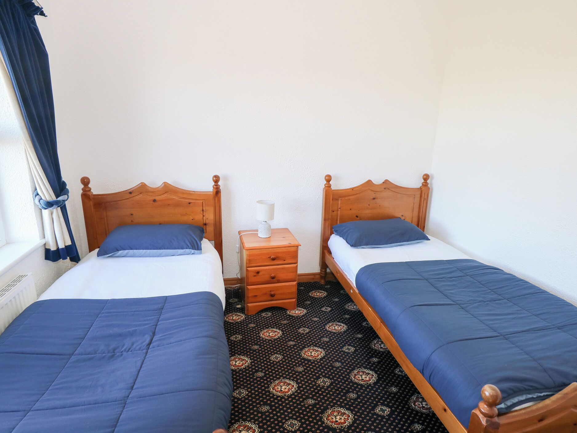 A room with two wooden beds, each with a blue bedspread and pillow, a small wooden nightstand with a lamp, and patterned carpet.