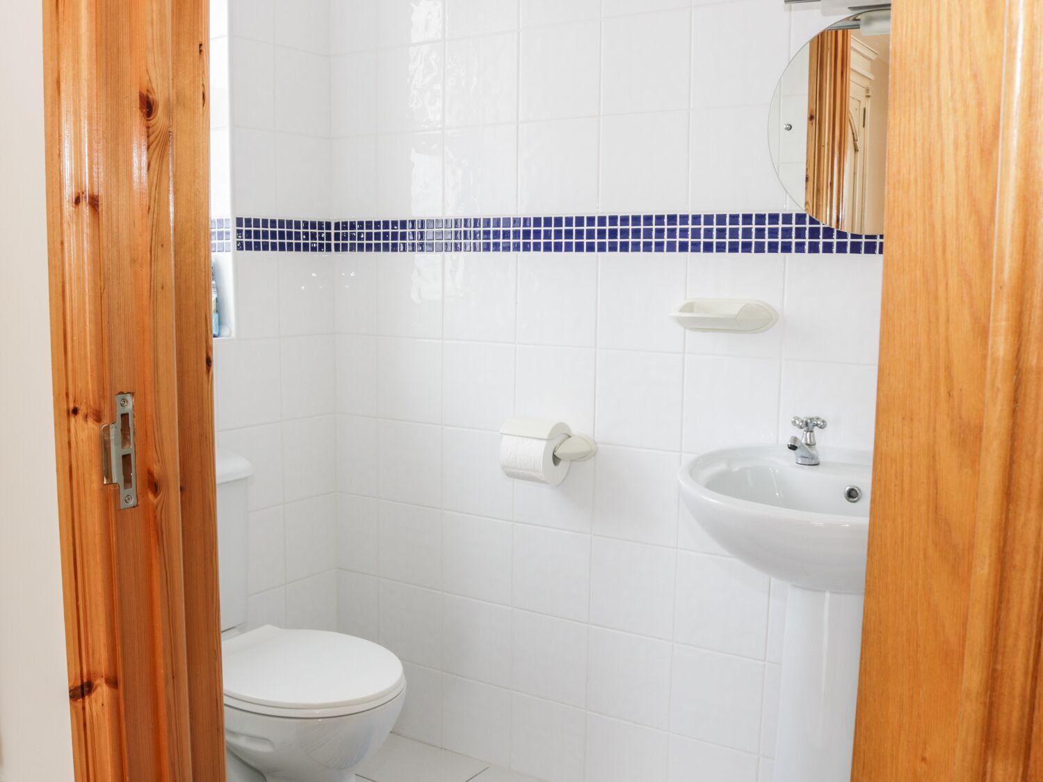 A bathroom with white tiles, a blue-tiled shower area, a white toilet, and a sink with a mirror above it.