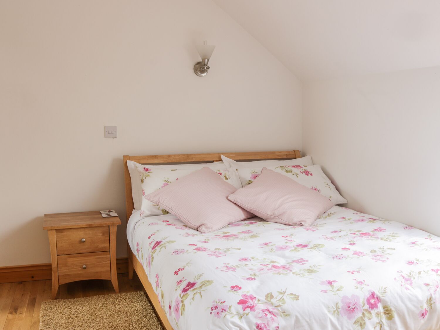 A bedroom with a wooden bed frame, a nightstand with a drawer, a wall-mounted light fixture, and a floral-patterned bedspread.