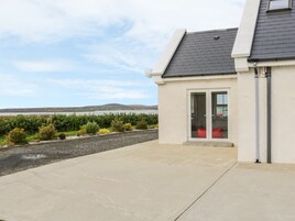 Una casa moderna con un gran ventanal, un patio de hormigón y vistas a un lago y colinas al fondo.