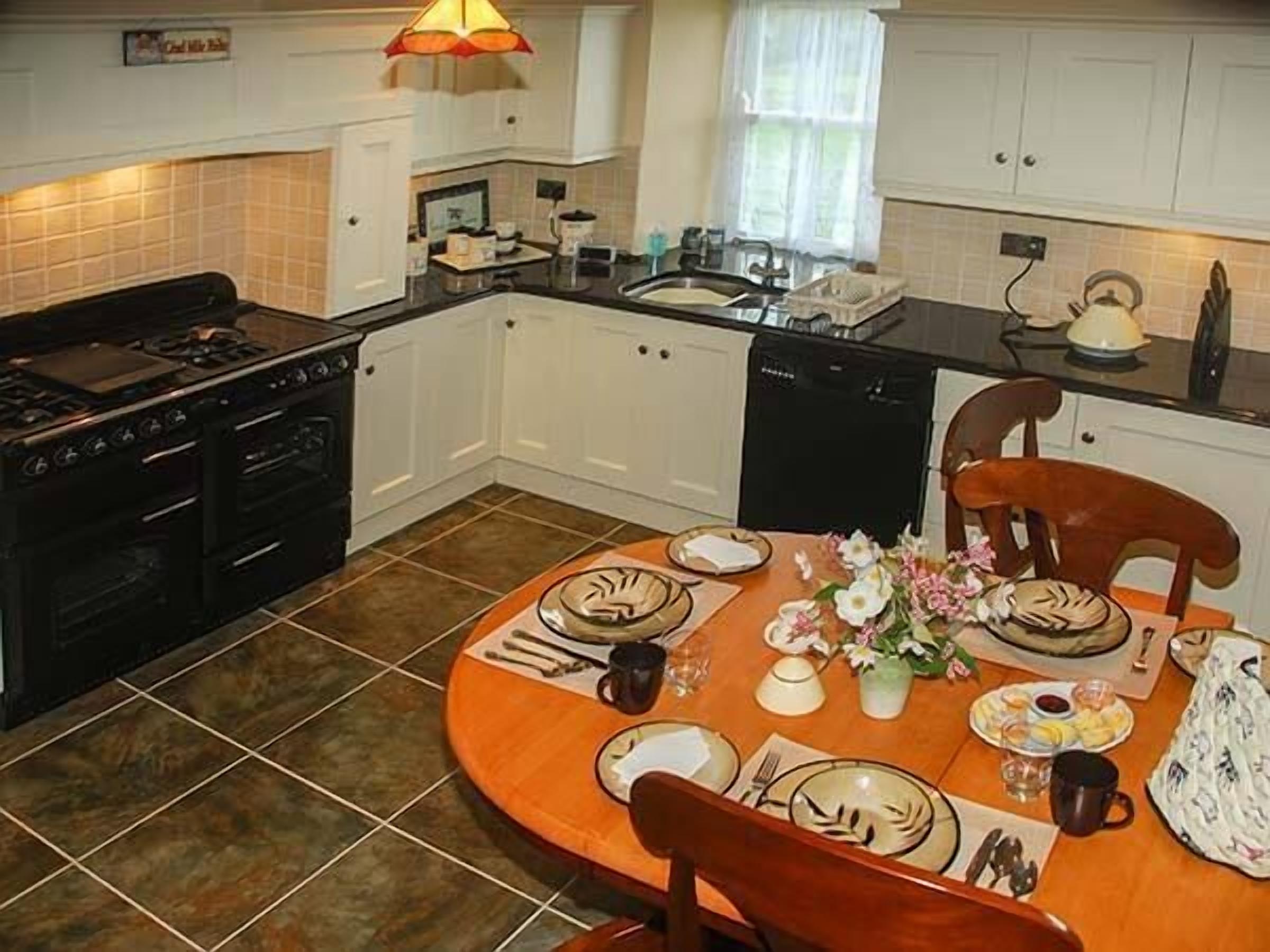 A modern kitchen with white cabinets, a black stove, and a dining table set for four with plates, cutlery, and a vase of flowers.