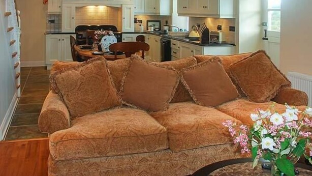 A living room with a brown sofa, a dining area with a table and chairs, and a kitchen with white cabinets. - Nellie's Farmhouse (Bantry)
