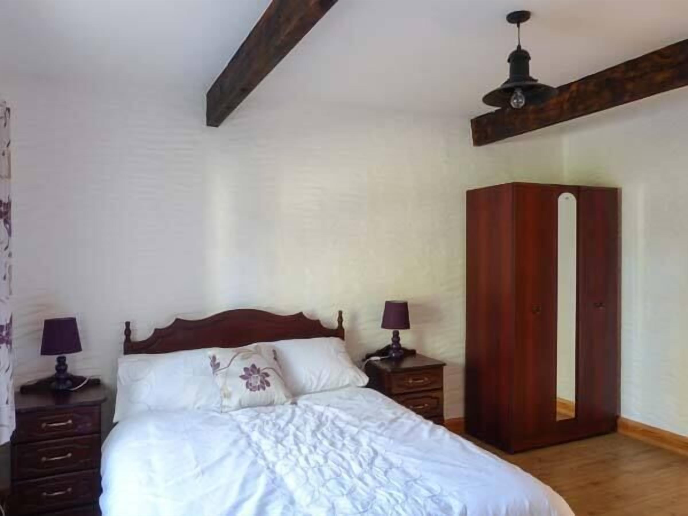 A bedroom with a wooden bed frame, white bedding, two bedside tables with lamps, and a wooden wardrobe.