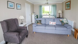 Un salon avec un canapé gris, un fauteuil marron, une petite table avec une lampe et une fenêtre avec des rideaux.