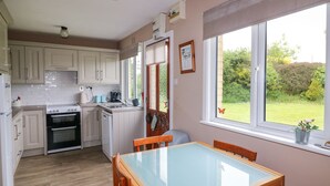 Une cuisine avec des meubles blancs, une cuisinière et un évier. On y trouve une table à manger avec des chaises et une fenêtre donnant sur le jardin.