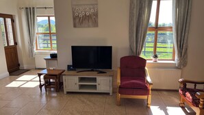 Sala de estar con televisor sobre un mueble bajo, un sillón rojo, una silla de madera y vistas al verdor a través de las ventanas.
