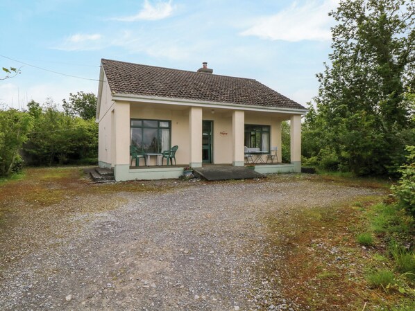 A single-story house with a tiled roof, a porch with two chairs, and a gravel driveway.