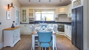 Une cuisine avec des meubles blancs, une table à manger avec des chaises bleues, un réfrigérateur et une lucarne.