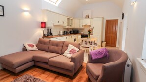 A compact living space with a sofa, armchair, and a small dining table. A kitchen is visible in the background with a window above the sink. - Laharn Cottage (Killorglin)