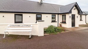 A two-story building with a flat roof, a white exterior, and a black door. There is a bench in front of the building, and a small garden area with plants and flowers. - Laharn Cottage (Killorglin)