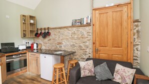 A compact kitchen with a stone wall, wooden cabinets, and a built-in oven. There is a small island with stools and a couch with floral cushions. - Swallow's Nest (Tacumshane)
