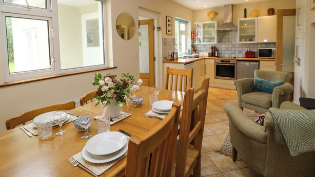 A dining area with a wooden table set for four, a kitchen in the background, and a cozy armchair with a throw blanket. - Rowantree (Killarney)