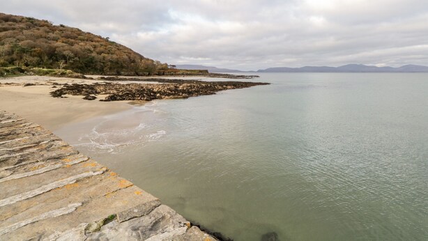 A coastal landscape with a rocky shoreline, a sandy beach, and a stone pier extending into calm waters. - At the Crossroads (Louisburgh)