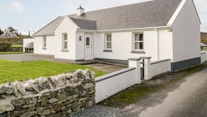 A white building with a stone wall in front, a gravel driveway, and a grassy area. - At the Crossroads (Louisburgh)